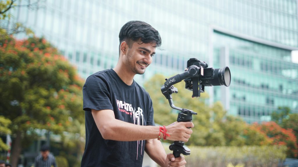 Smiling young videographer using a gimbal outdoors. Urban setting and greenery in the background.